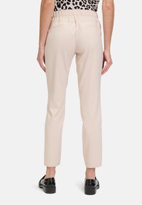 Beżowe spodnie o dopasowanym kroju i elastycznym pasie. Widok z tyłu pokazuje dwie kieszenie i zwężane nogawki, zestawione z błyszczącymi czarnymi butami.