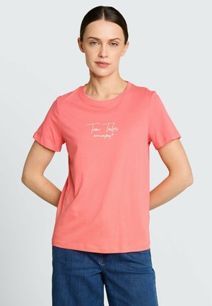 Frau mit zurückgebundenem dunklem Haar, die ein korallenrosa T-Shirt mit weißem Text und blaue Jeans trägt, steht mit den Händen hinter dem Rücken.