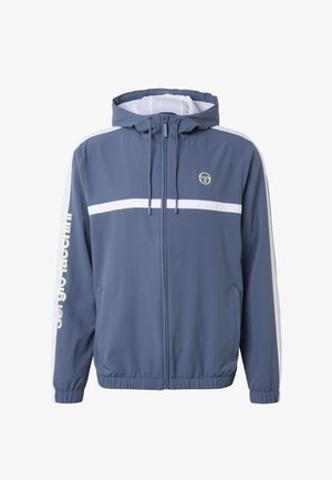 Blauwe zip-up jas met capuchon, witte horizontale streep over de borst, witte strepen op de mouwen en de tekst "Sergio Tacchini" op de linkerarm.