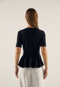 Černý peplum top s krátkými rukávy, kulatým výstřihem a staženým pasem. Hladký materiál se strukturovaným tvarem, v kombinaci s bílými kalhotami.