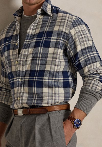 Mand iført blå og hvid ternet skjorte over grå sweater, grå bukser, brun læderbælte og ur med blå urskive og brun rem.