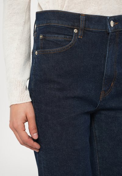 Σκούρο μπλε τζιν με ραμμένες ραφές, πέντε τσέπες και κούμπωμα με κουμπί. Το ύφασμα έχει μια λεία υφή με διακριτική ξεθώριαση.
