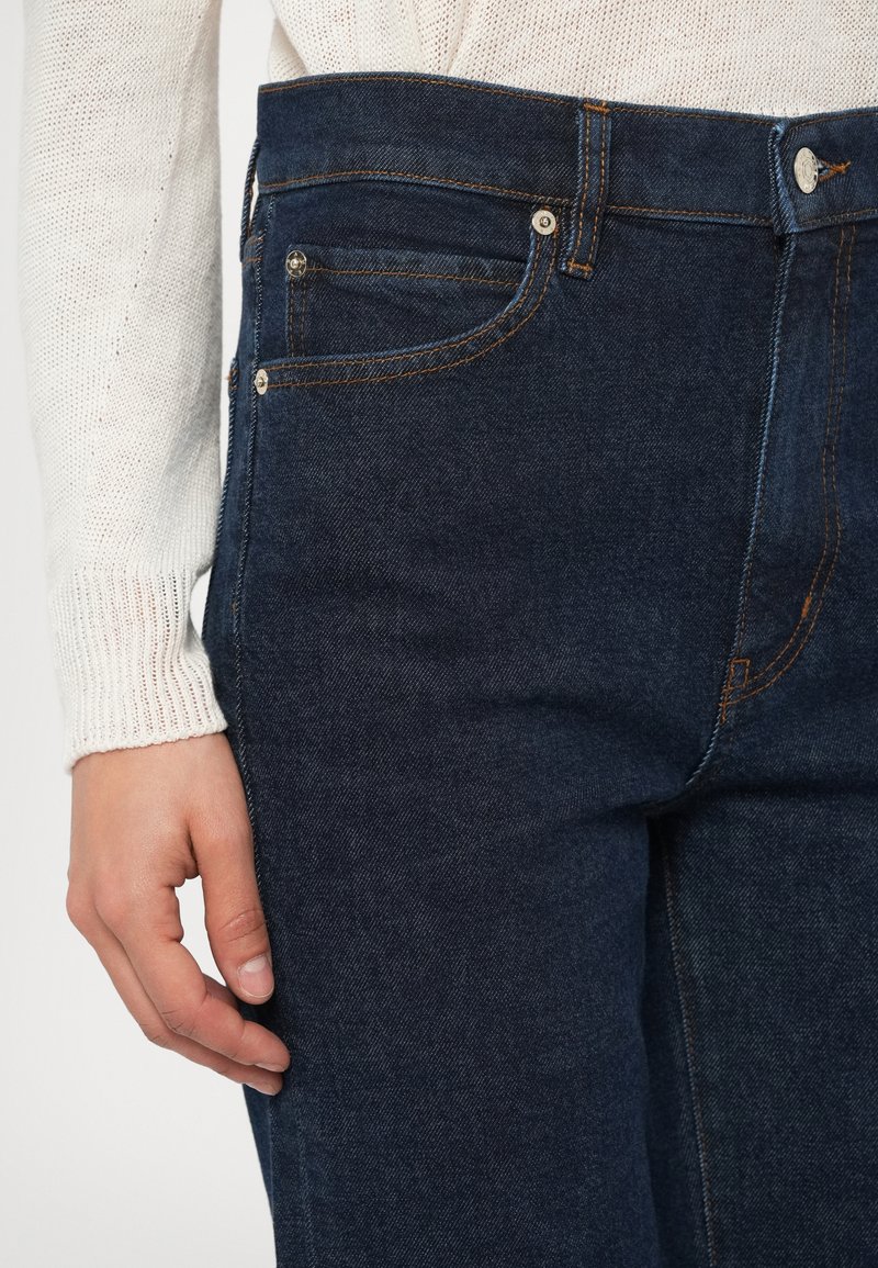 Dunkelblaue Jeans aus Denim mit gesteppter Naht, fünf Taschen und einem Knopfverschluss. Der Stoff hat eine glatte Textur mit dezentem Verblassen.