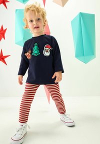 Kék, hosszú ujjú ing ünnepi motívumokkal (rénszarvas, fa, Télapó) kombinálva piros-fehér csíkos leggings-szel és fehér sportcipőkkel.