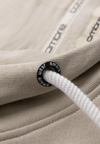 Detail béžové látky s černým kovovým okem a bílým šňůrkou. Na štítku tkaniny a oku je zobrazeno branding "Ombre".