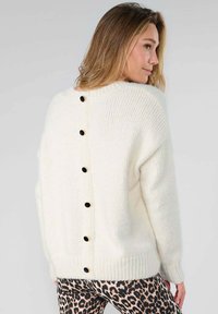 Crème trui met een geribbelde textuur, voorzien van een knoopsluiting aan de achterkant en lange mouwen. Gecombineerd met een broek in panterprint.
