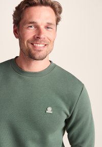 Smiling man with blond hair wearing a green TOG24 sweatshirt against a plain light background.