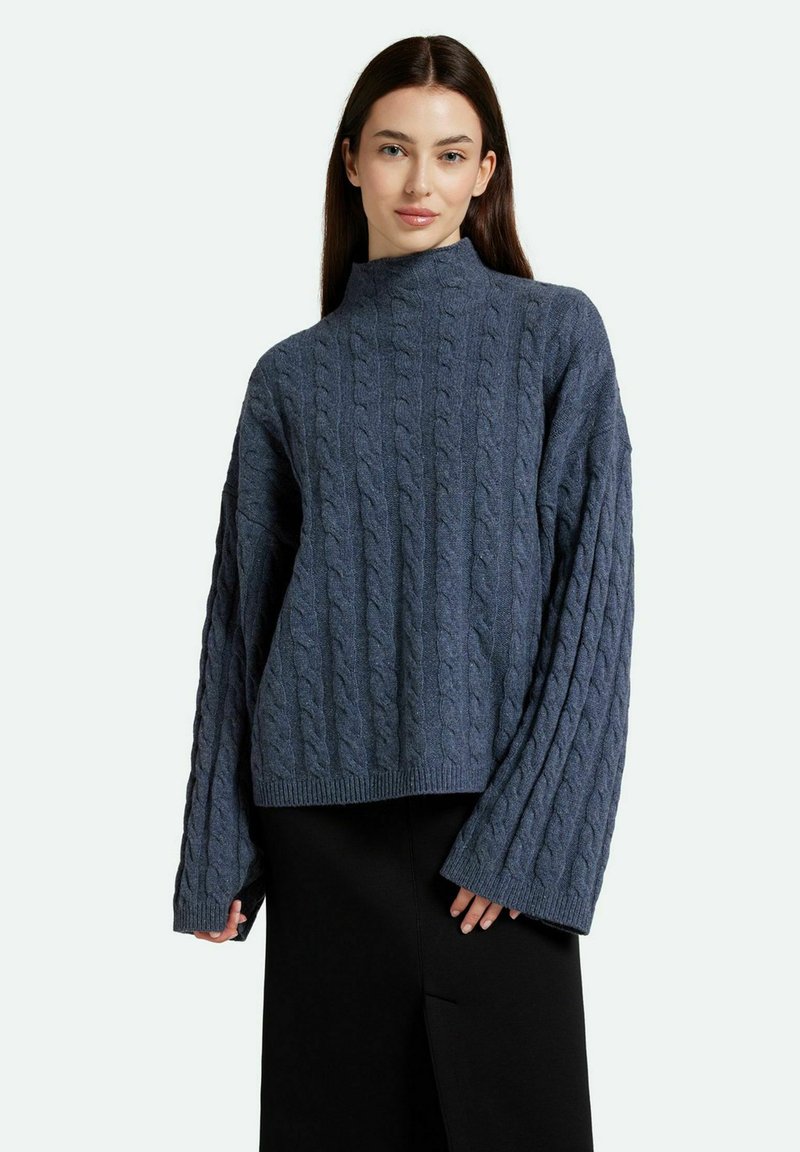 Pull en tricot torsadé bleu marine avec col montant, manches larges et ourlet côtelé. Le motif texturé ajoute de la profondeur au design.