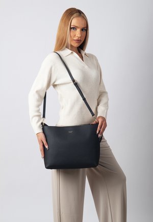 Svart läder tote-väska med en pebbled yta och guldfärgade detaljer. Har en justerbar axelrem och ett rymligt inre, hålls av en modell.