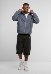 Hettegrå hettegenser med glidelås, kombinert med svarte denimshorts. Hvite joggesko og ankelsokker fullfører antrekket. Enkelt design, uformell passform.