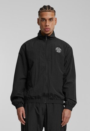 Black windbreaker jacket with a zip front, high collar, elastic cuffs, and hem, featuring a circular logo on the left chest. Smooth texture.