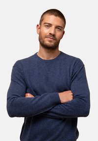 Młody mężczyzna z krótkimi włosami i brodą, ubrany w niebieski sweter, stoi z rękami skrzyżowanymi, pewnie patrząc w obiektyw.