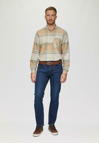 Chemise à boutons en beige avec un motif à carreaux ; accents bleus et verts. Jean foncé ajusté et baskets brunes avec des lacets foncés. Ceinture marron.