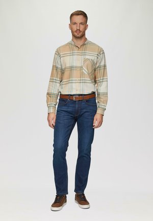 Chemise à boutons en beige avec un motif à carreaux ; accents bleus et verts. Jean foncé ajusté et baskets brunes avec des lacets foncés. Ceinture marron.