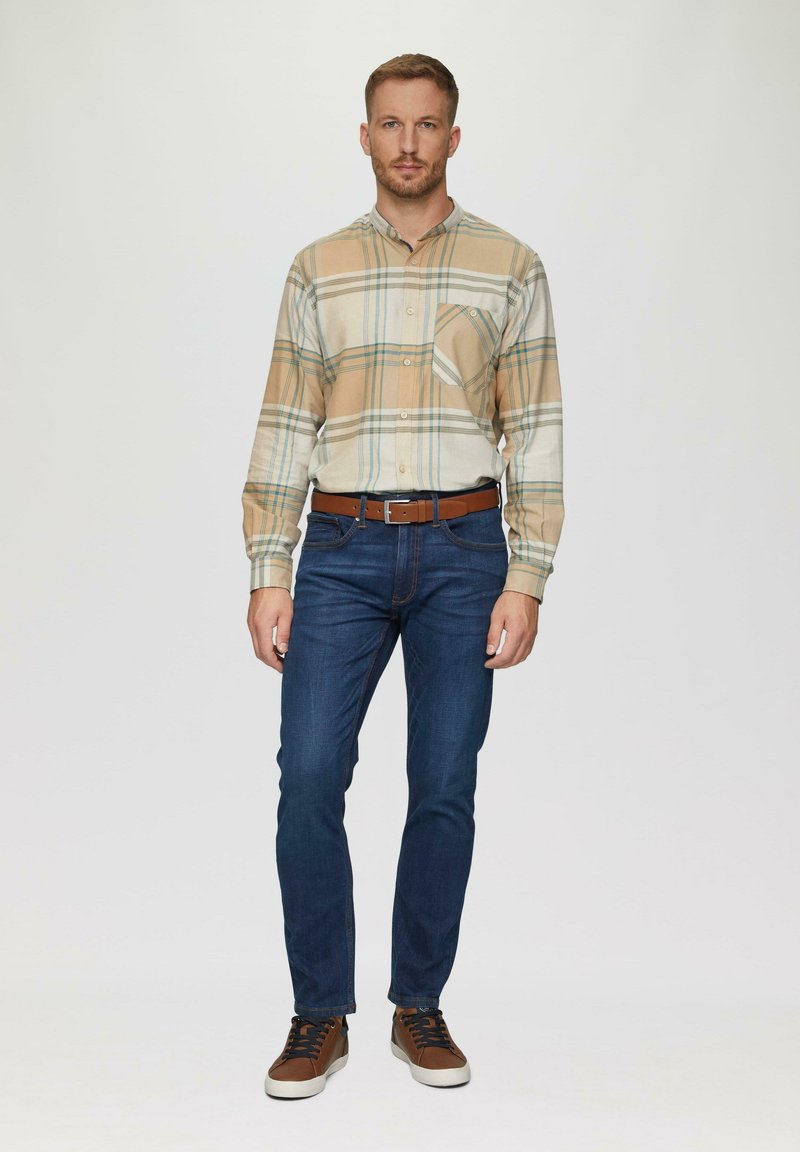 Chemise à boutons en beige avec un motif à carreaux ; accents bleus et verts. Jean foncé ajusté et baskets brunes avec des lacets foncés. Ceinture marron.