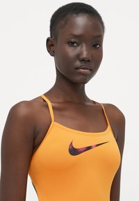Mlada žena s kratkom kosom nosi narančasti Nike top s tankim trakama na bijeloj pozadini.