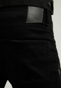 Jeans de mezclilla negra con una textura suave, que presentan un parche de cuero negro en la cinturilla y un diseño de bolsillo trasero cosido.