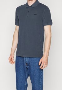 Polo z krótkim rękawem w ciemnoszarym kolorze z bawełny, z zapięciem na guziki, detalem logo na klatce piersiowej i klasycznym kołnierzykiem. Połączony z niebieskimi dżinsami.