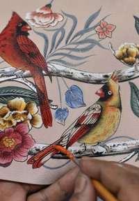 Illustratie van twee kleurrijke vogels op gestructureerd beige leer, omringd door bloemen en bladeren; kunstenaar voegt details toe met een penseel.