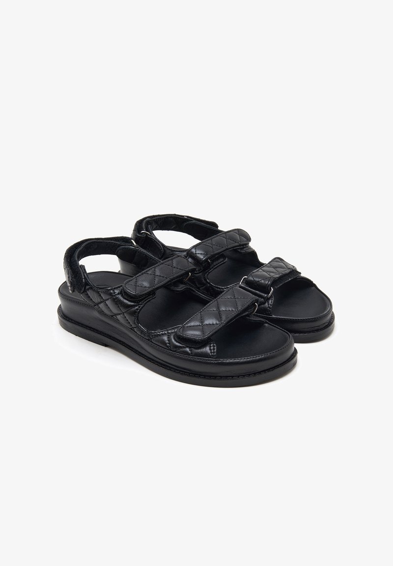 Sandales en cuir noir avec brides matelassées et fermetures Velcro réglables, dotées d'une semelle plate et d'un design à bout ouvert.