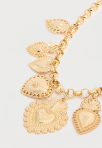 Kuld käevõru kaunistatud südame- ja lehekujuliste ripatsitega, mis on kaunistatud keerukate lille- ja tekstuuriliste mustritega valgel taustal.