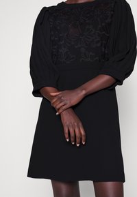 Černé šaty s krajkovým topem s květinovými vzory, nabíranými balónovými rukávy a přiléhavým pasem. Jemná textura s hladkým povrchem.