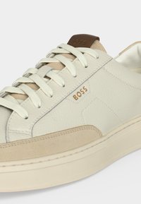 Λευκό δερμάτινο sneaker χαμηλής κοπής με μπεζ σουέντ λεπτομέρειες, λευκά κορδόνια, χοντρή σόλα και ανάγλυφο λογότυπο "BOSS" στο πλάι και στη γλώσσα.