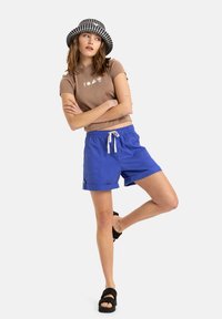 Brun kortärmad skjorta med vitt grafiskt tryck, blå shorts med snörning och fickor, svartvitt rutigt bucket-hatt, tjocka svarta sandaler.
