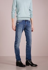 Svetle modrý svetr, vybledlé modré džíny s mírnými oděrkami a černé kotníkové boty s detailním přezkou. Jednoduché pozadí.