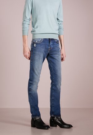 Lichtblauwe trui, vervaagde blauwe spijkerbroek met lichte slijtage, en zwarte enkellaarsjes met een gesp-detail. Effen achtergrond.