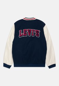 Námořnický modrý varsity kabát s krémovými rukávy, se velkým vyšitým logem "LEVI'S" v červené a bílé barvě. Pruhovaný límec a manžety.
