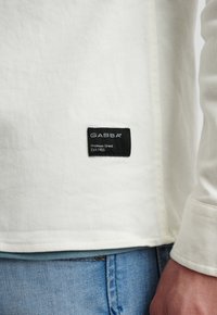 T-shirt bianco a maniche lunghe con finitura testurizzata, caratterizzato da un'etichetta in tessuto nera cucita sul lato sinistro inferiore. Abbinato a jeans blu.