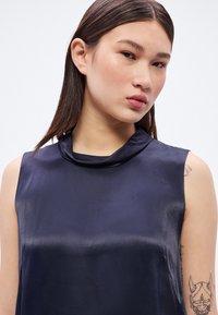 Námornícky modrý saténový top bez rukávov s dizajnom rolovaného výstrihu, hladkou textúrou a pohodlným strihom, s viditeľným tetovaním na pravej ruke.