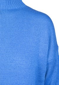 Prim-plan cu un pulover tricotat albastru intens, evidențiind textura fină cu nervuri și cusăturile de pe umăr și partea superioară a mânecii.