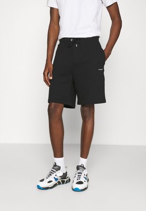 Mężczyzna ubrany w czarne szorty ze ściągaczem, białe skarpetki oraz czarno-biało-niebieskie sportowe buty, stoi na jednolitym tle.