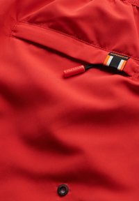 Tela de chaqueta roja con una textura suave. Presenta un bolsillo con cremallera, detalle de botón negro y una etiqueta con rayas y logotipo.