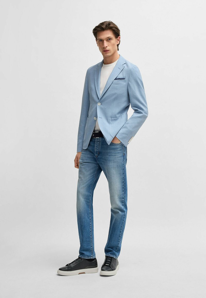 Light blue blazer with textured fabric, single button closure, paired with faded denim jeans and black sneakers. White t-shirt underneath.