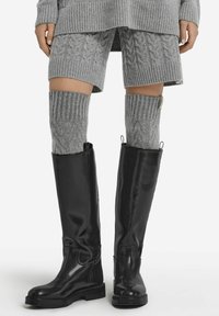 Czarne, skórzane botki sięgające do kolan o błyszczącej powierzchni, w zestawieniu z szarymi, sztruksowymi skarpetami sięgającymi do ud oraz szarym, prążkowanym strojem, ukazującym warstwowe faktury.