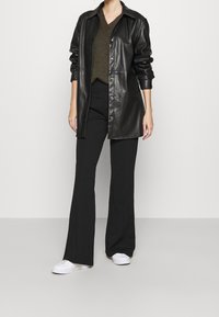 Svart läder skjortjacka med knäppning framtill, bärs över en olivgrön V-ringad tröja och svarta utsvängda byxor, kompletterat med vita sneakers.