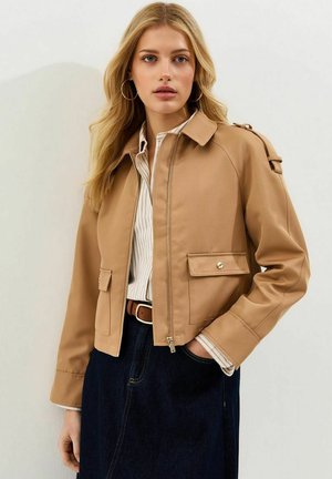 Blondynka w dużych kołowych kolczykach, opalona krótka kurtka na pasku w paski, ciemna spódnica z brązowym paskiem, stoi na jednolitym tle.