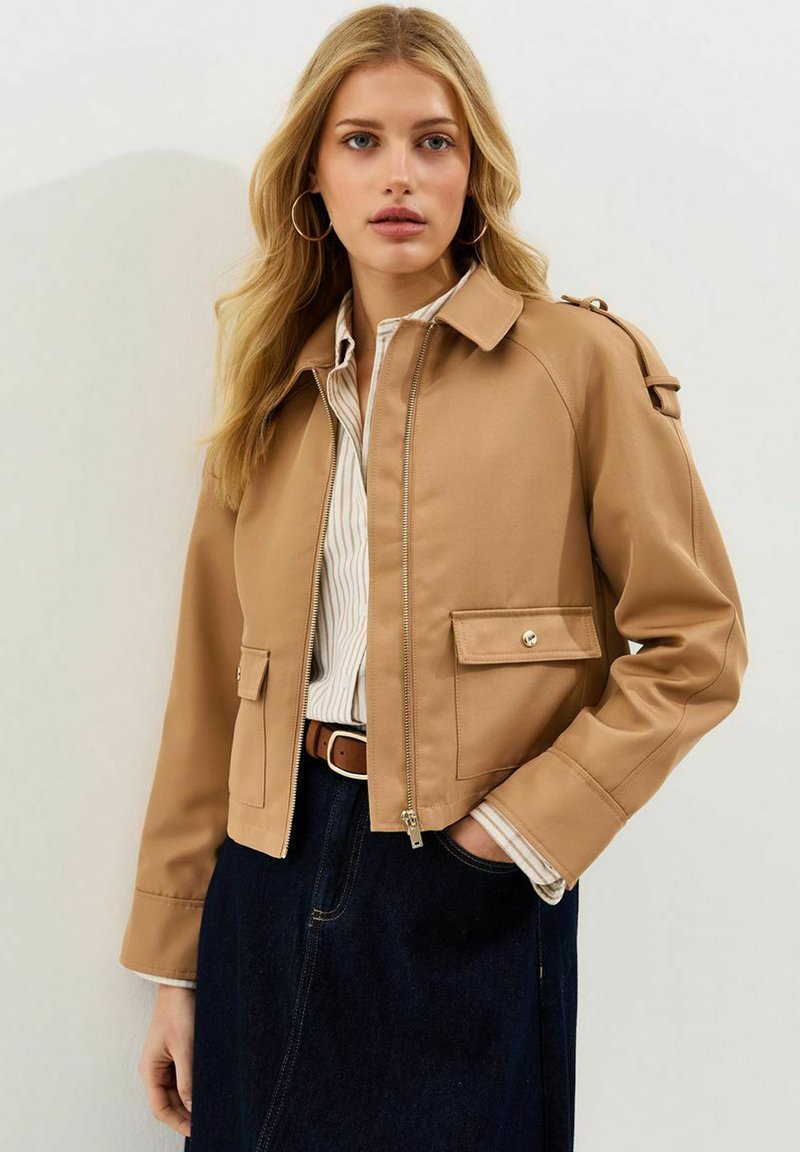 Mujer rubia con grandes pendientes de aro, chaqueta corta color bronceado sobre camisa de rayas, falda oscura con cinturón marrón, de pie contra un fondo liso.