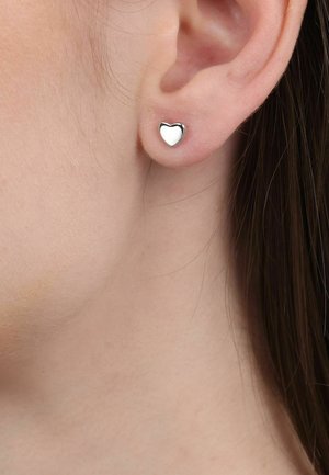 Close-up van het oor van een persoon met een kleine, gepolijste zilveren oorknop in de vorm van een hartje.