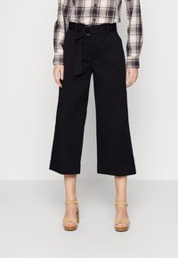 Czarne, szerokie spodnie o kroju 7/8 z pasem i detalem na szlufce. Połączone z jasnymi sandałami na obcasie. Na górze widoczna koszula w kratę.