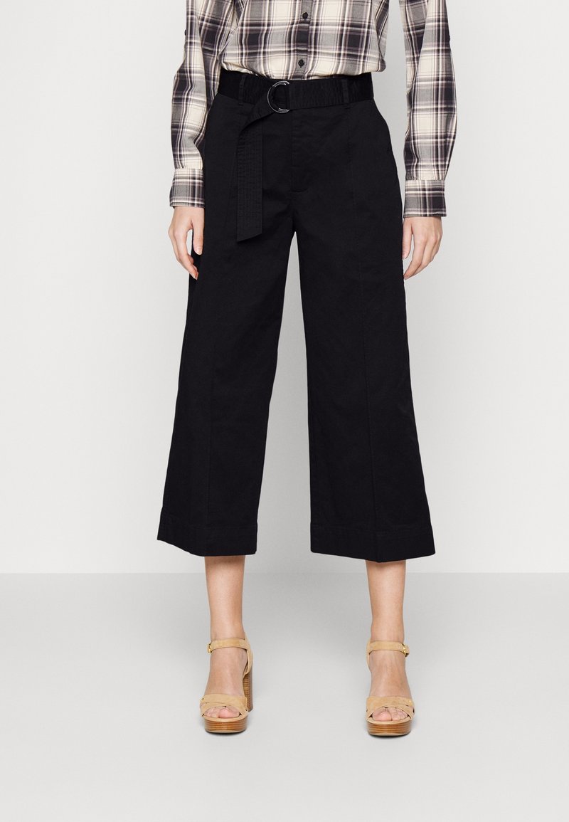 Czarne, szerokie spodnie o kroju 7/8 z pasem i detalem na szlufce. Połączone z jasnymi sandałami na obcasie. Na górze widoczna koszula w kratę.