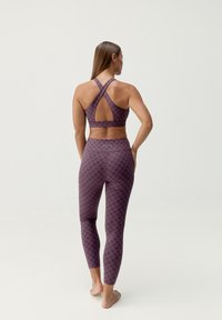 Sujetador deportivo morado con diseño de espalda cruzada, combinado con leggings de cintura alta con un patrón negro repetido. Tejido suave y elástico.