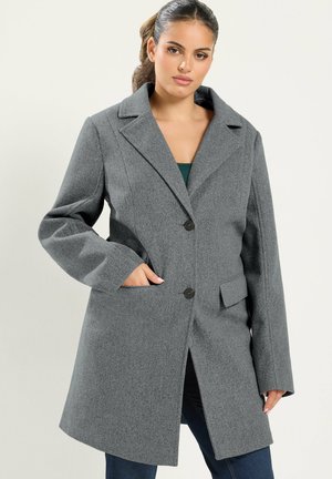 Femme portant un manteau en laine gris avec deux boutons et des poches, debout devant un fond clair uni avec une main dans une poche.