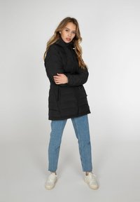 Sort dunjakke med høj krave og lynlås. Har sidelommer og en quiltet tekstur. Parret med lyseblå jeans og hvide sneakers.