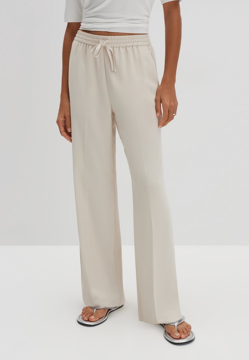 Personne portant un pantalon beige à jambes larges avec taille à cordon, un top blanc court et des sandales flip-flop argentées sur fond uni.