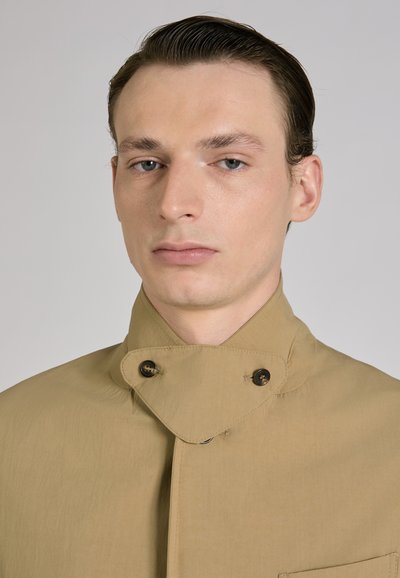 Jonge man met naar achteren geveegd haar, gekleed in een beige jas met hoge kraag, dichtgeknoopt met twee donkere knopen, tegen een effen achtergrond.