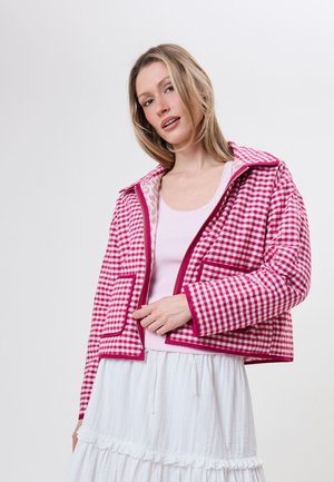 Vrouw draagt een roze-wit geruite jas over een lichtroze top en een witte geplooide rok tegen een effen achtergrond.